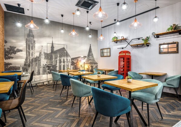 Interior de restaurante com um mural de uma catedral, mesas e cadeiras de madeira, luminárias pendentes e uma cabine telefônica vermelha.