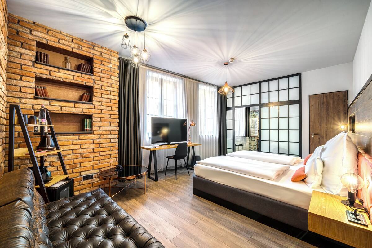 Una habitación de hotel moderna con pared de ladrillo, una cama grande, un escritorio con televisor y un sofá de cuero.