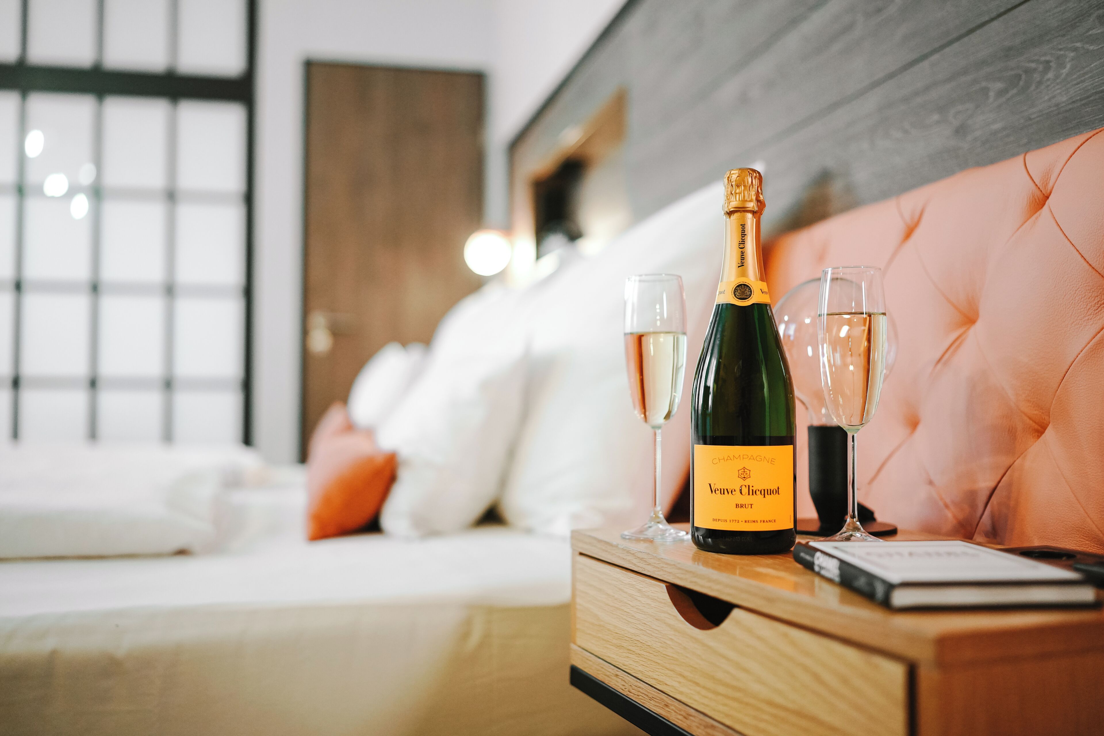 A bedroom with a bed, a bedside table with a bottle of champagne and two glasses, and a book.