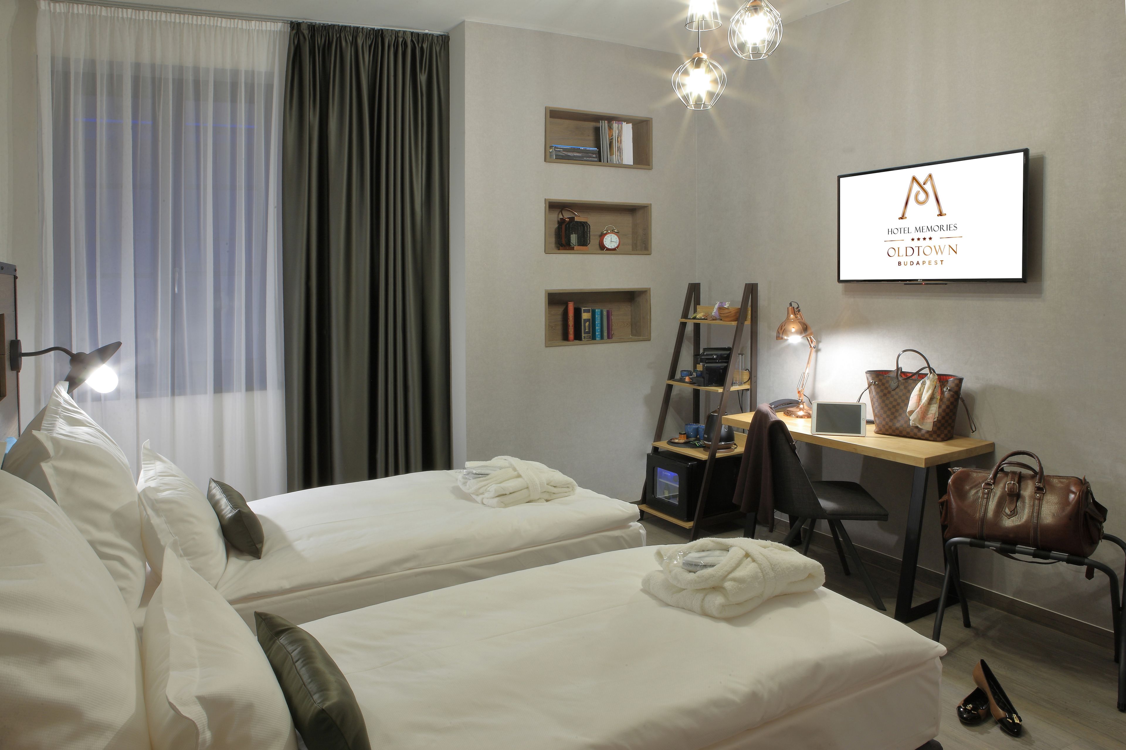 Ein Hotelzimmer mit zwei Betten, einem Schreibtisch, einem Stuhl, einer Lampe, einer Handtasche und einem an der Wand befestigten Fernseher, der den Namen des Hotels anzeigt.