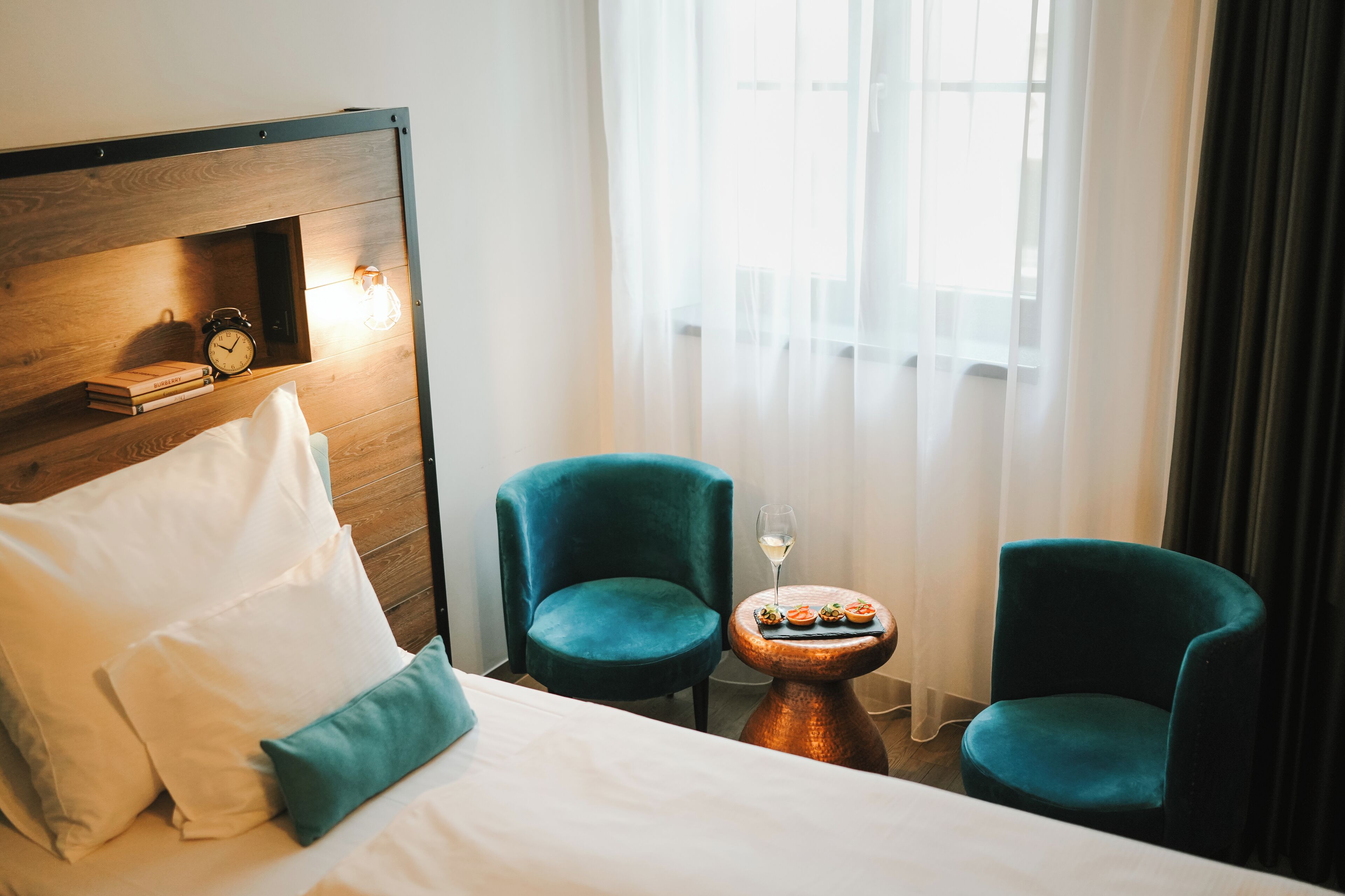 A hotel room with a bed, two chairs, a small table with a glass of wine, and a window with curtains.