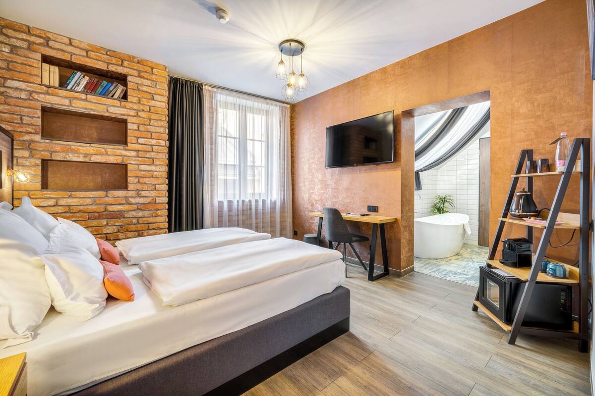 Una habitación de hotel moderna con una pared de ladrillo decorativa, un televisor de pantalla plana, un escritorio con una silla, una bañera y una estantería.