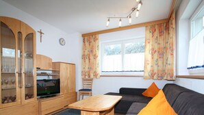 Tunnelmallinen olohuone, jossa on puinen kaappi, televisio, oransseilla tyynyillä varustettu sohva ja ruokapöytä.