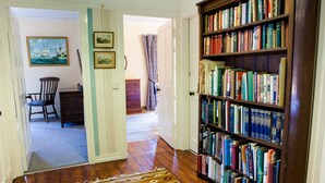 Un pasillo con piso de madera, un tapete estampado, una estantería llena de libros, una silla y una puerta que da a otra habitación.