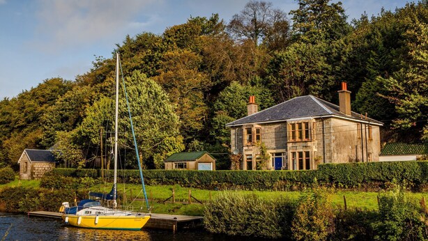 Una casa junto a un río, con un muelle y un velero.