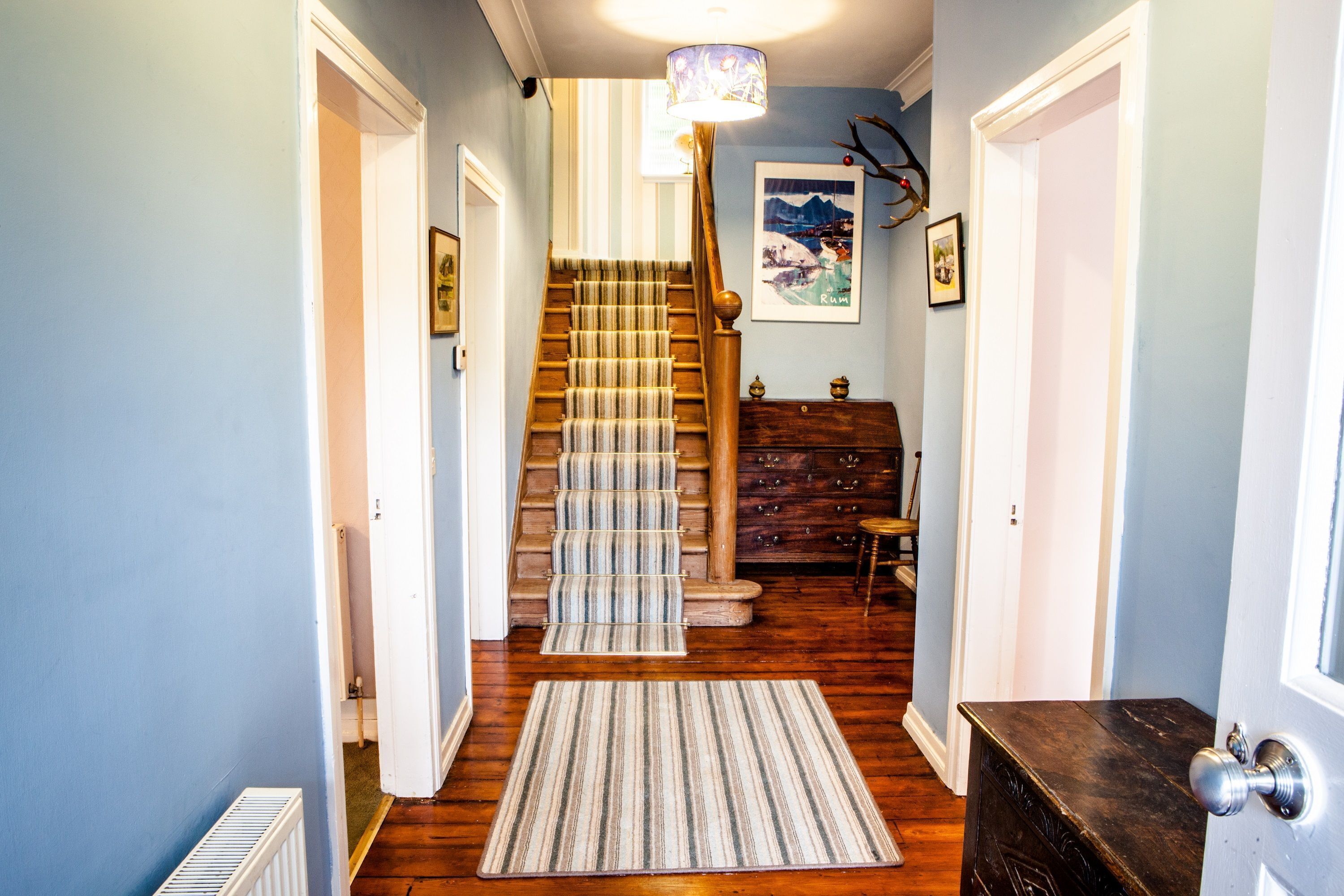 Un pasillo con una escalera de madera, un radiador, un felpudo a rayas y una puerta con picaporte plateado.