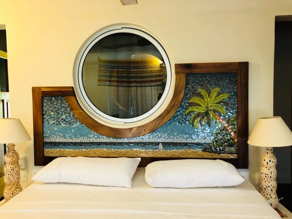 Una habitación de hotel con una cama, dos lámparas de noche, un espejo redondo y un muro de mosaico con una palmera.