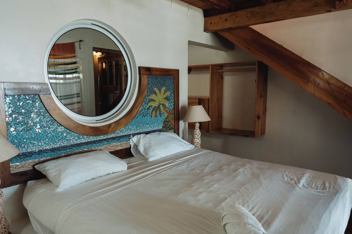 Quarto com cama, espelho redondo, cabeceira de madeira, mesa de cabeceira e luminária.