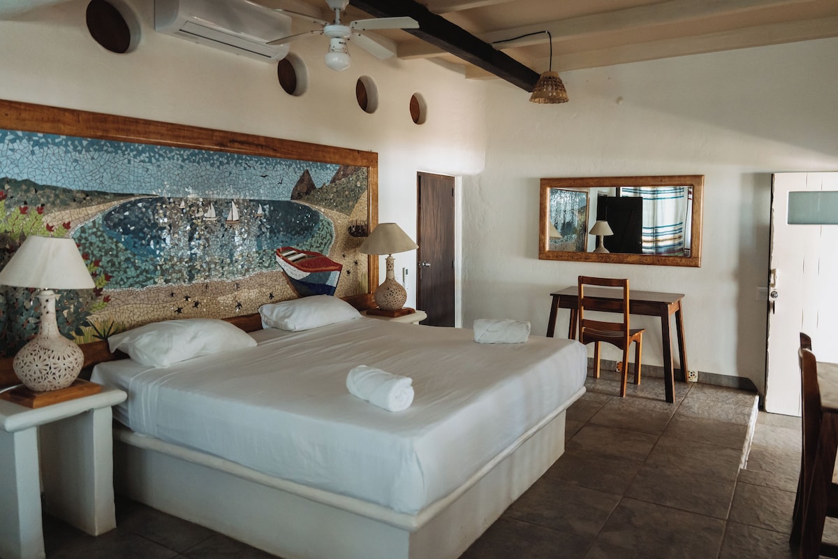 A hotel room with a large bed, a desk, a chair, a ceiling fan, and a mosaic wall.