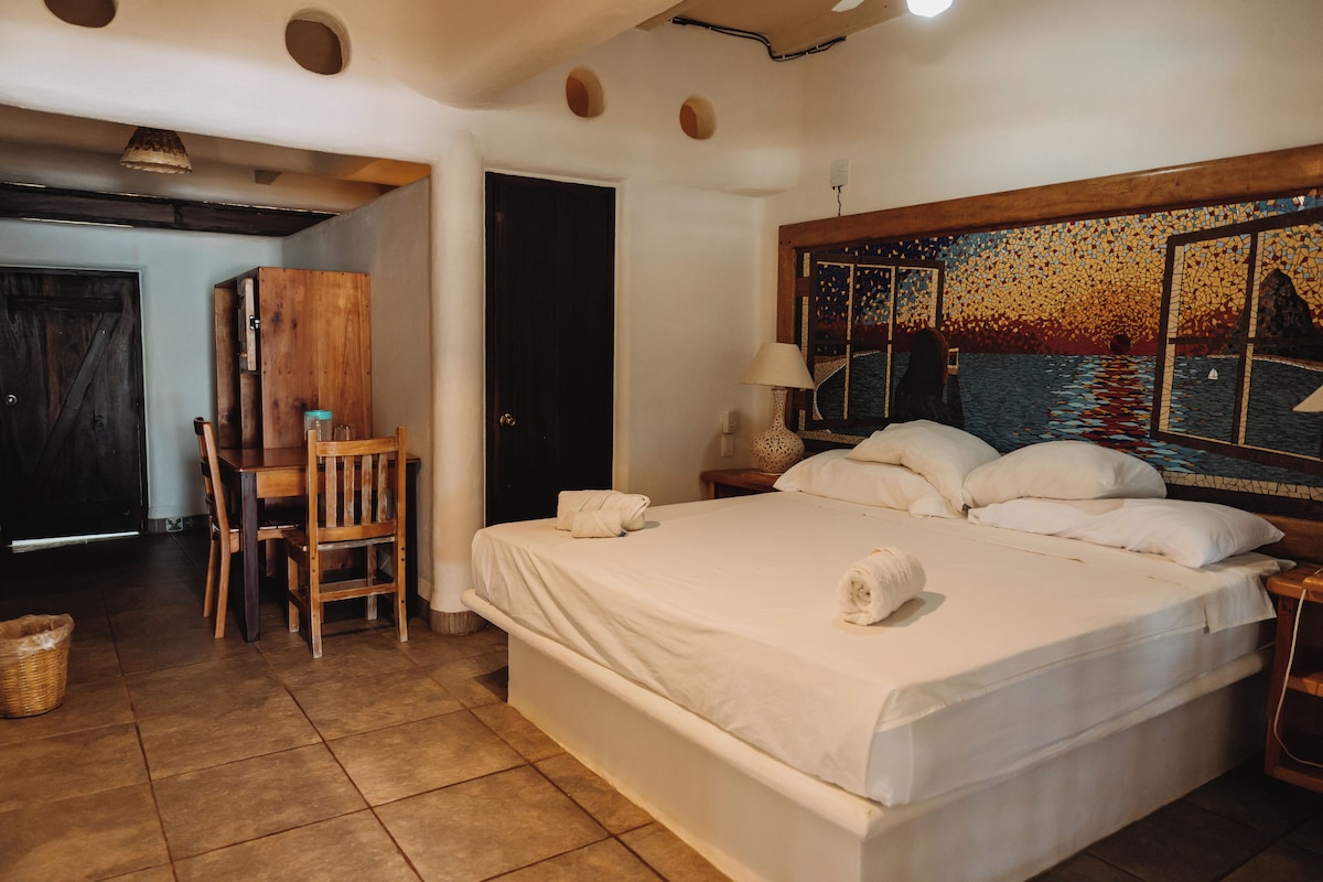 A hotel room with a large bed, a desk, a chair, a wooden wardrobe, and a mosaic artwork on the wall.