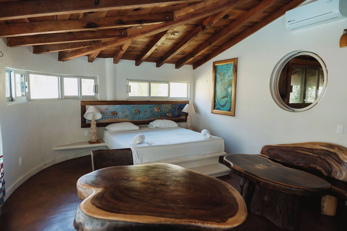 Um quarto com teto de madeira, uma cama com roupa de cama branca, um espelho redondo e um quadro na parede.