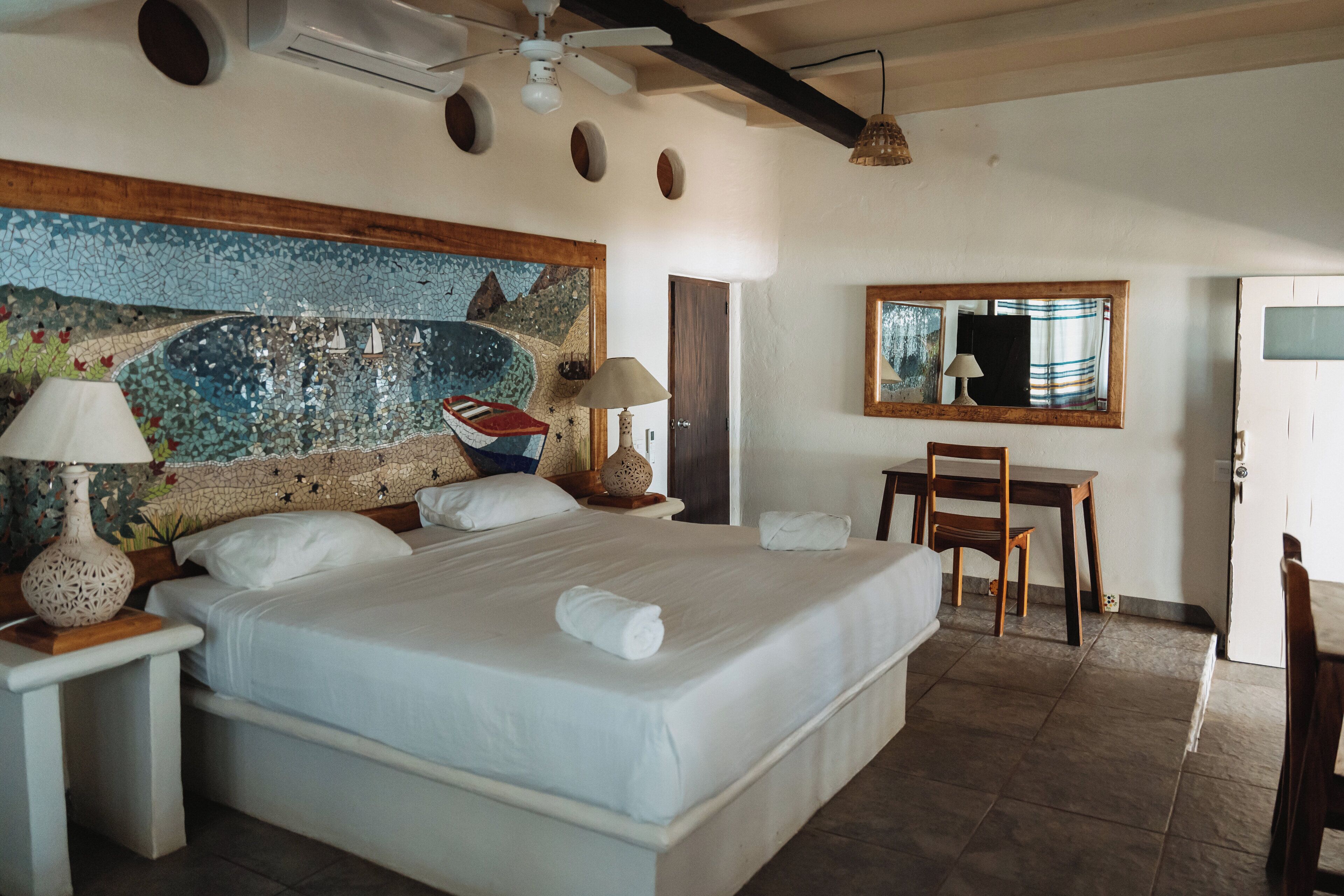 A hotel room with a large bed, a desk, a chair, a ceiling fan, and a mosaic wall.