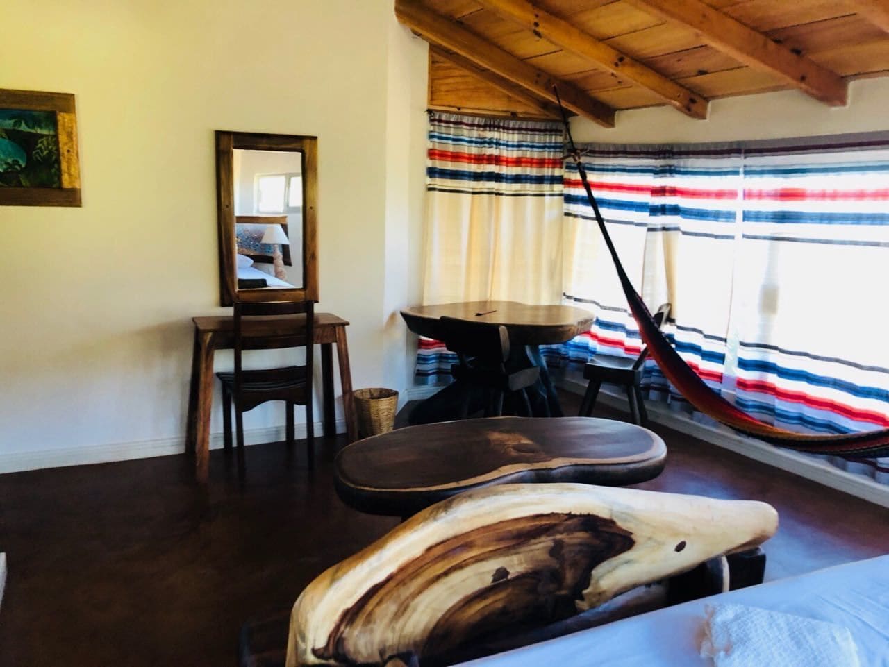 A room with a wooden ceiling, a dining table with chairs, a hammock, and a large mirror on the wall.