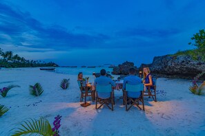 A group of people dining at a table on a sandy beach with a view of the ocean and boats in the distance. - SeVi Boutique Hotel (Matemwe)