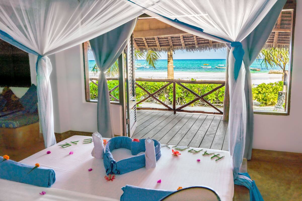 A room with a bed, a view of the beach, and a thatched roof structure.