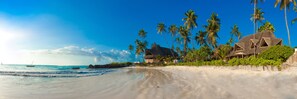 A beach with thatched-roof buildings, palm trees, and a clear blue sky. - SeVi Boutique Hotel (Matemwe)
