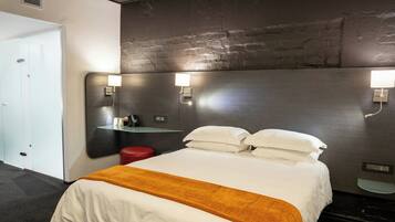 Une chambre d’hôtel avec un lit, un tabouret rouge, une étagère en verre et deux lampes fixées au mur.