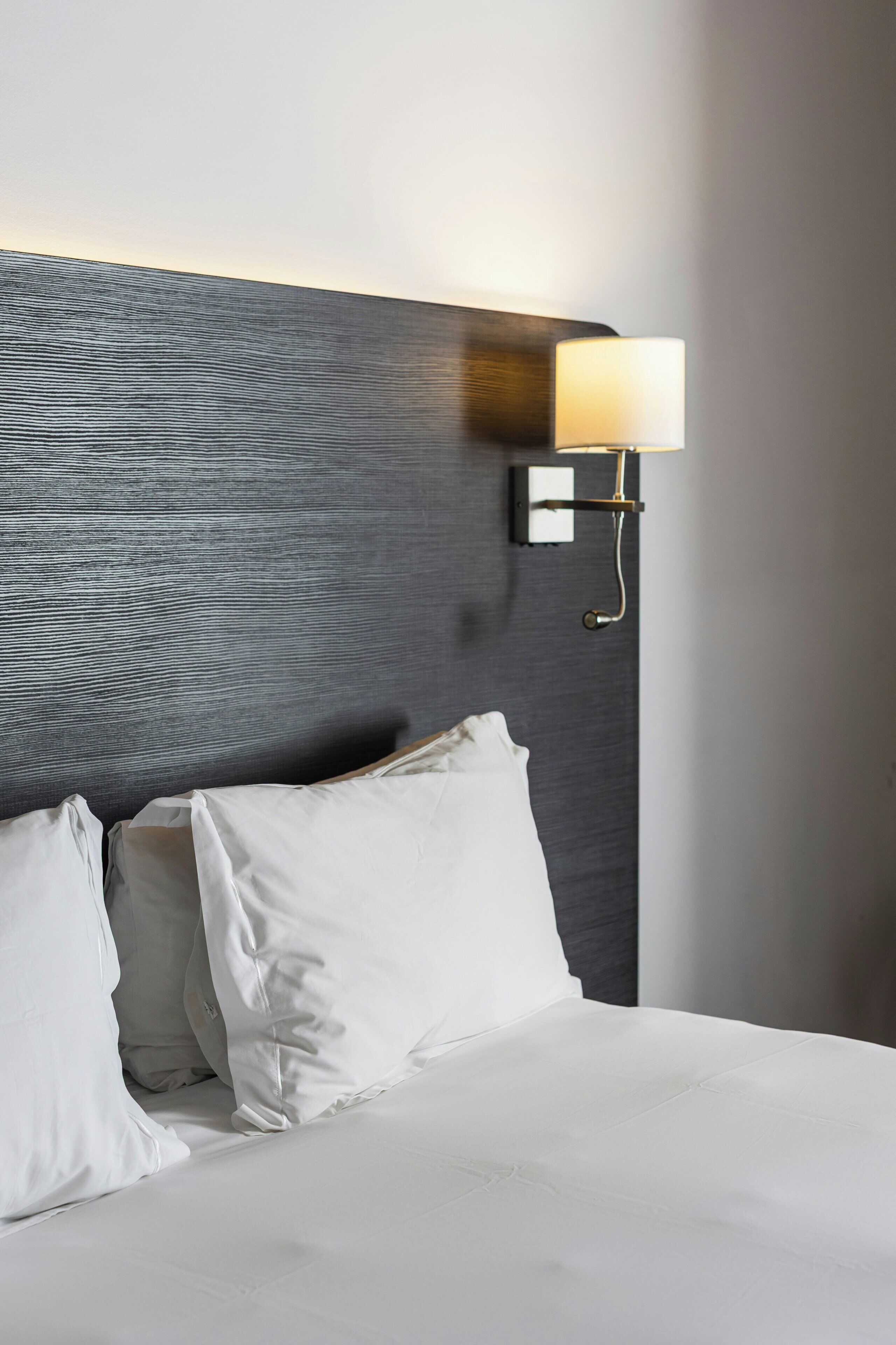 Habitación de hotel con cama, almohadas blancas, cabecera con textura y lámpara de pared.
