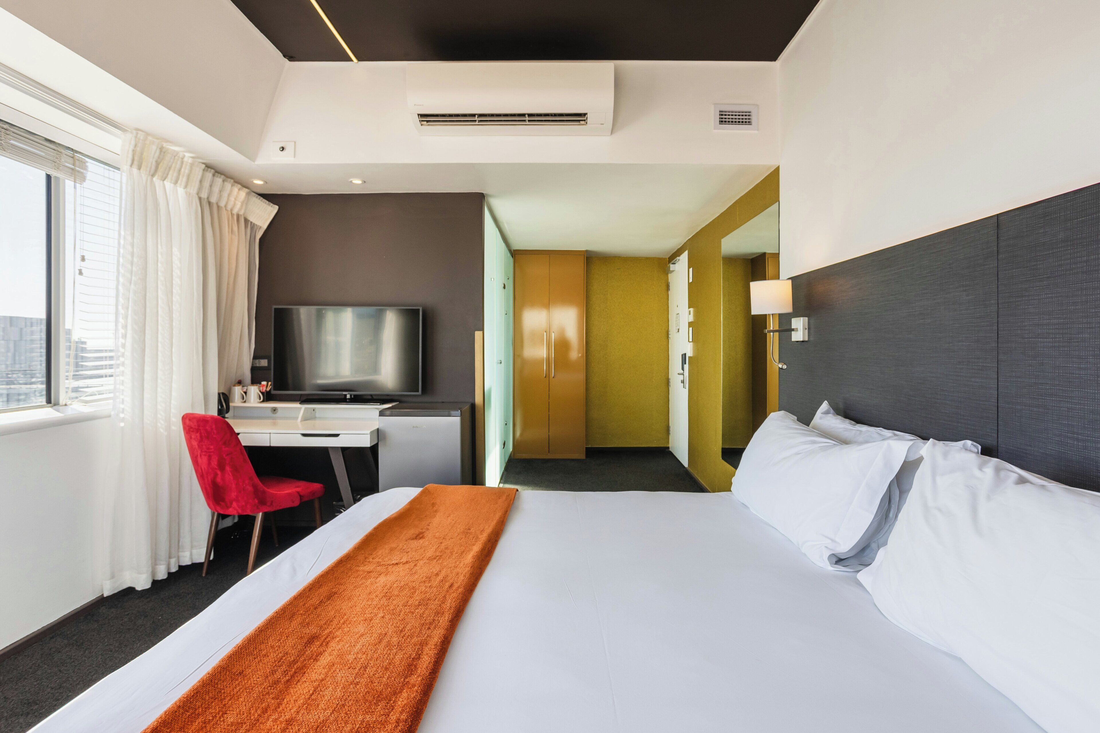 Habitación de hotel con cama, escritorio con televisor, una silla roja y vistas a la ciudad.