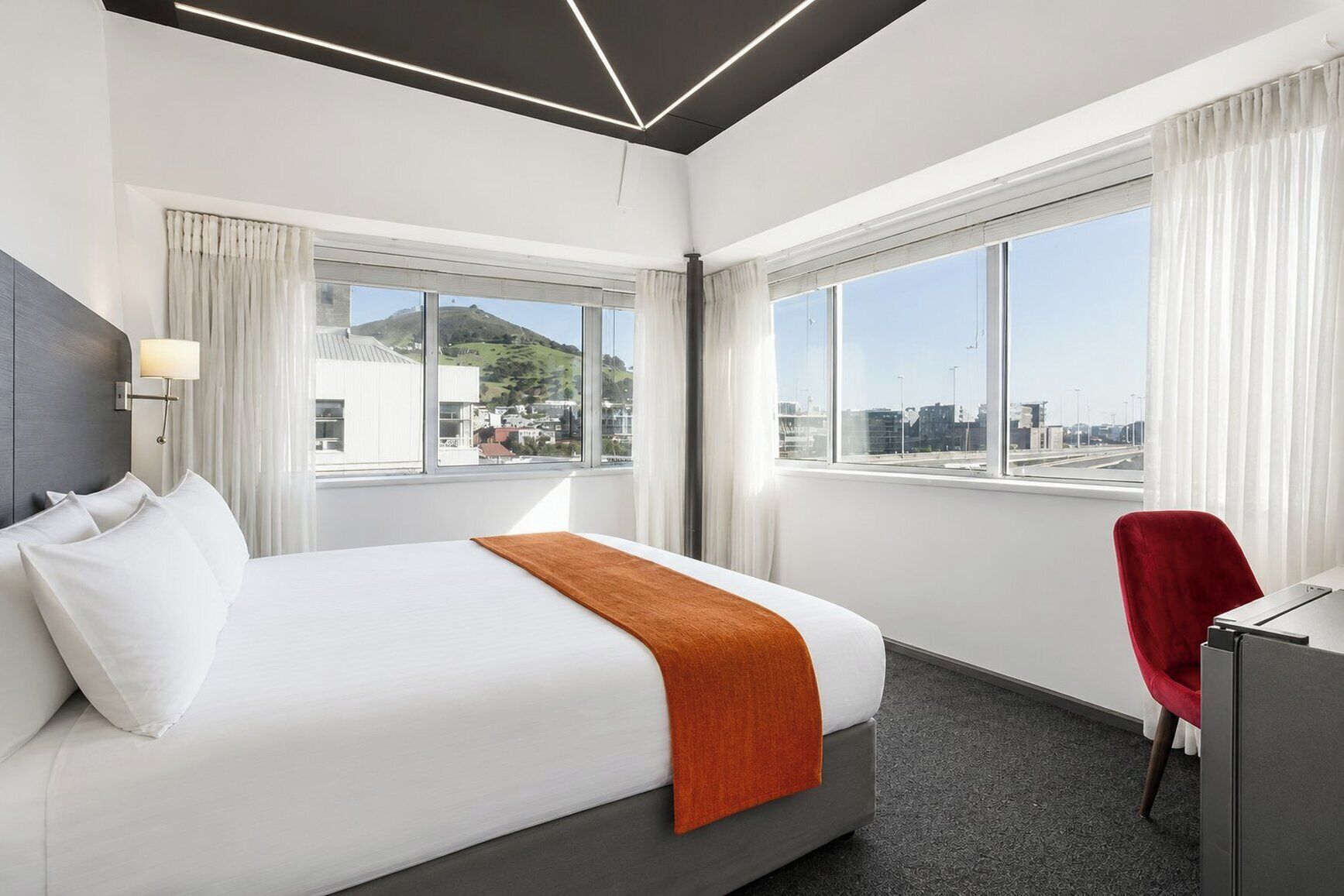 A hotel room with a large bed, a desk, and a chair. There are two windows with curtains, and a view of a cityscape and hills.