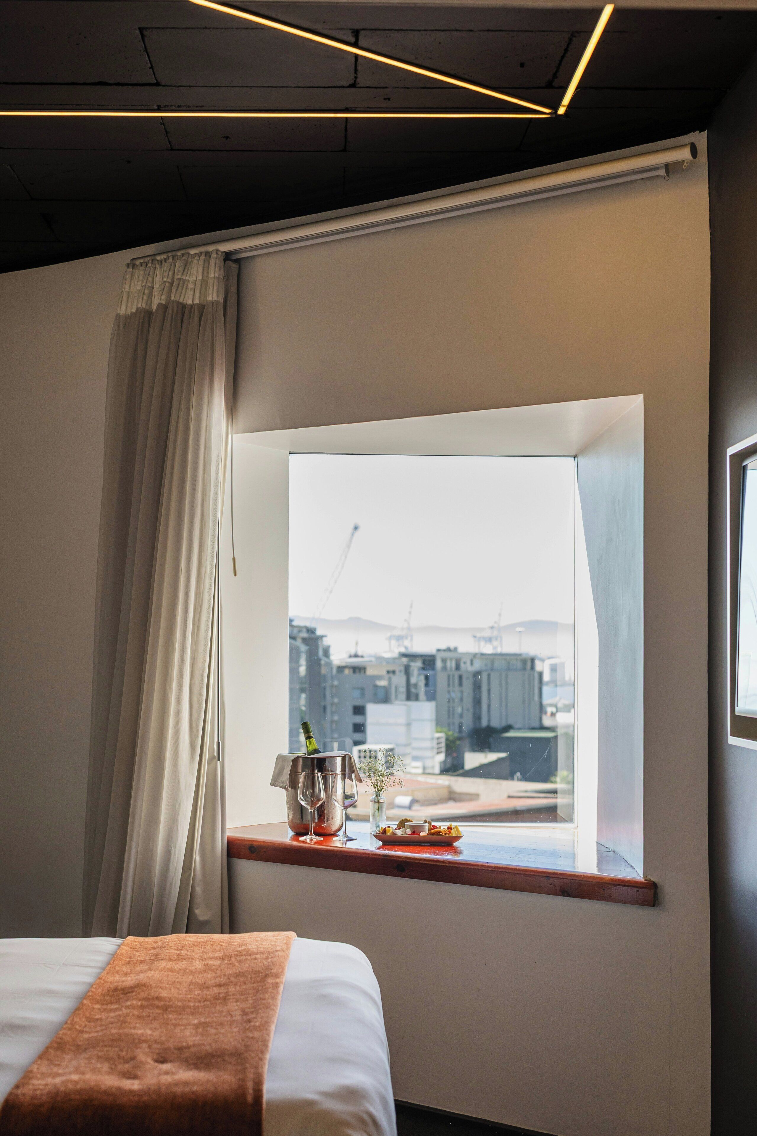 Une chambre d’hôtel avec un lit, une fenêtre donnant sur des immeubles, une table avec une bouteille, des verres et une assiette de nourriture.