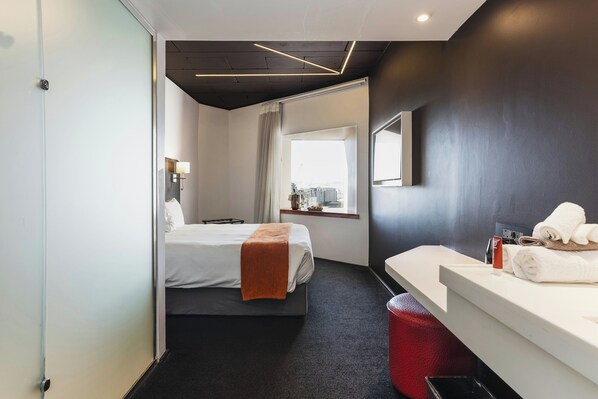 A hotel room with a bed, a bedside table with a mirror, a window with curtains, and a red trash bin. - ONOMO Hotel Waterfront (Cape Town)