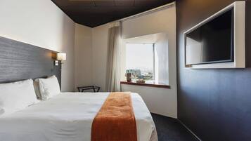 A hotel room with a bed, a TV mounted on the wall, a window with a view, and a ceiling with a geometric light fixture.