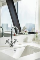 A modern bathroom with a large sink, chrome faucets, and a window providing a view of the cityscape.