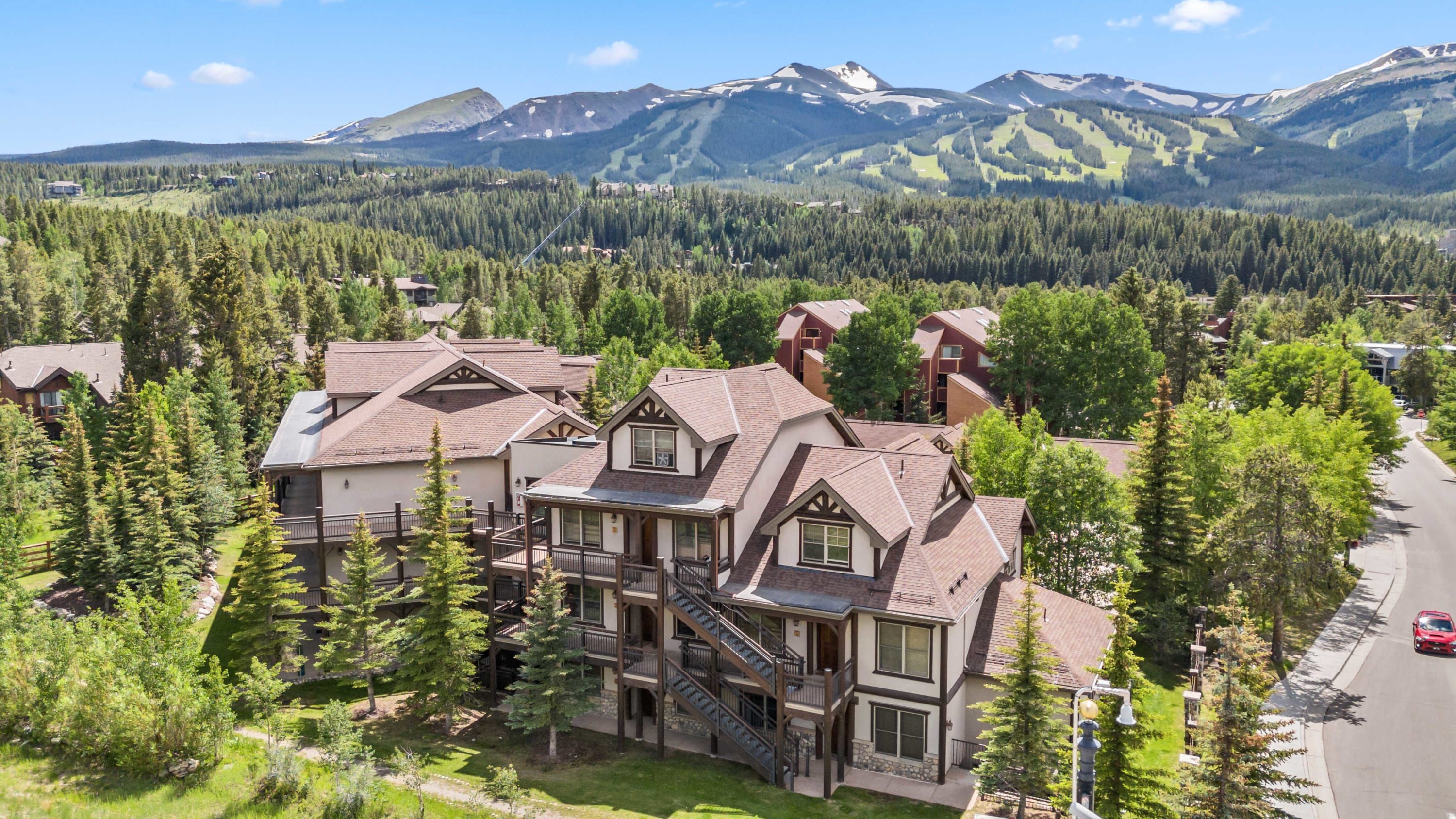 A multi-story residential building with a balcony, surrounded by trees and a mountain backdrop.