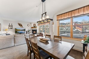 Ein gemütliches Wohnzimmer mit Kamin, Esstisch und großem Fenster mit Blick auf eine verschneite Landschaft.