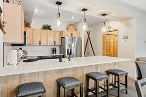 A modern kitchen with wooden cabinets, a stainless steel refrigerator, a white countertop, and three bar stools.