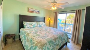 Un dormitorio con cama, ventilador de techo, mesita de noche, lámpara y ventana con cortinas.