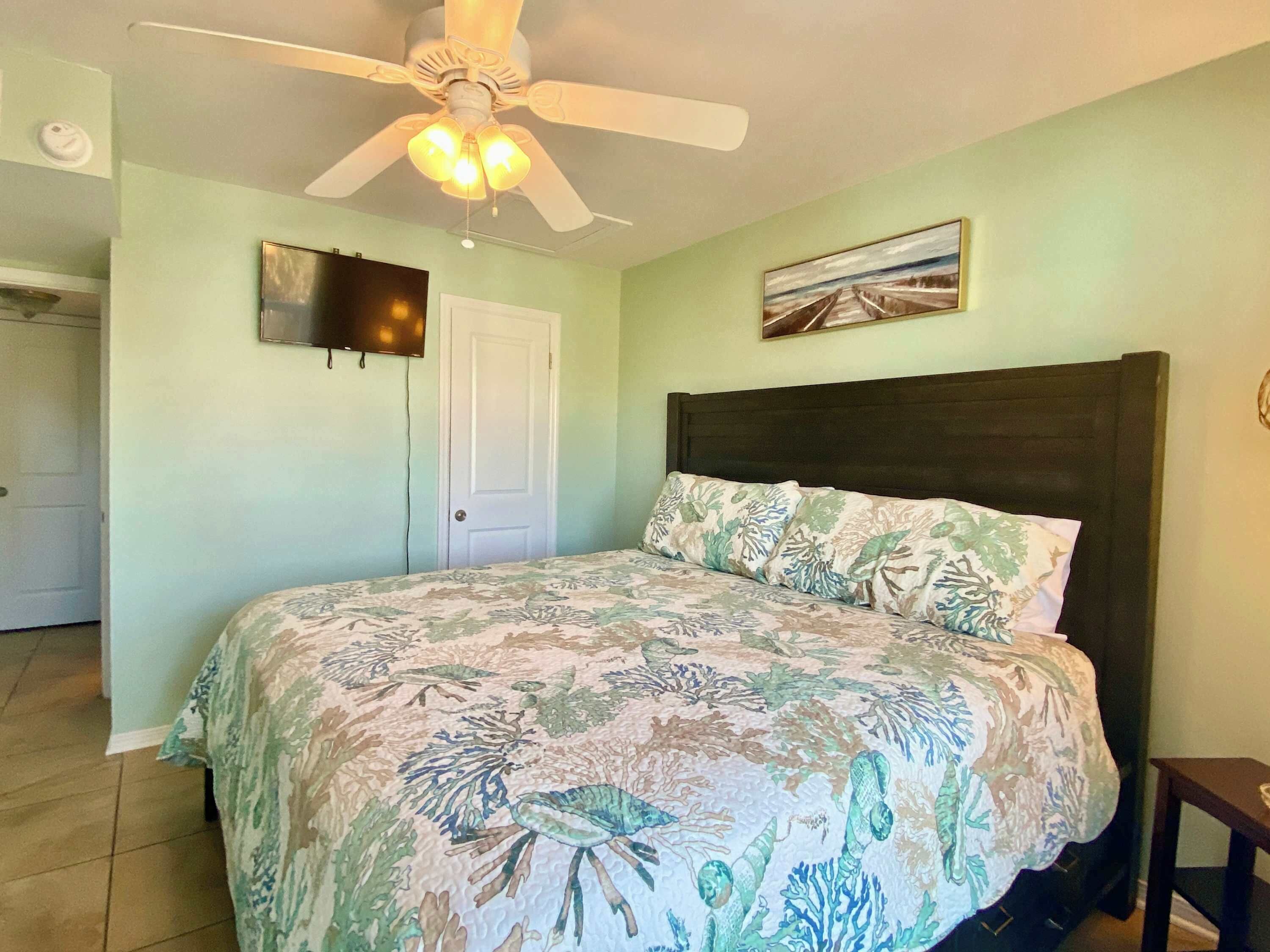 Une chambre à coucher avec un lit, un ventilateur de plafond, un téléviseur fixé au mur et une porte.