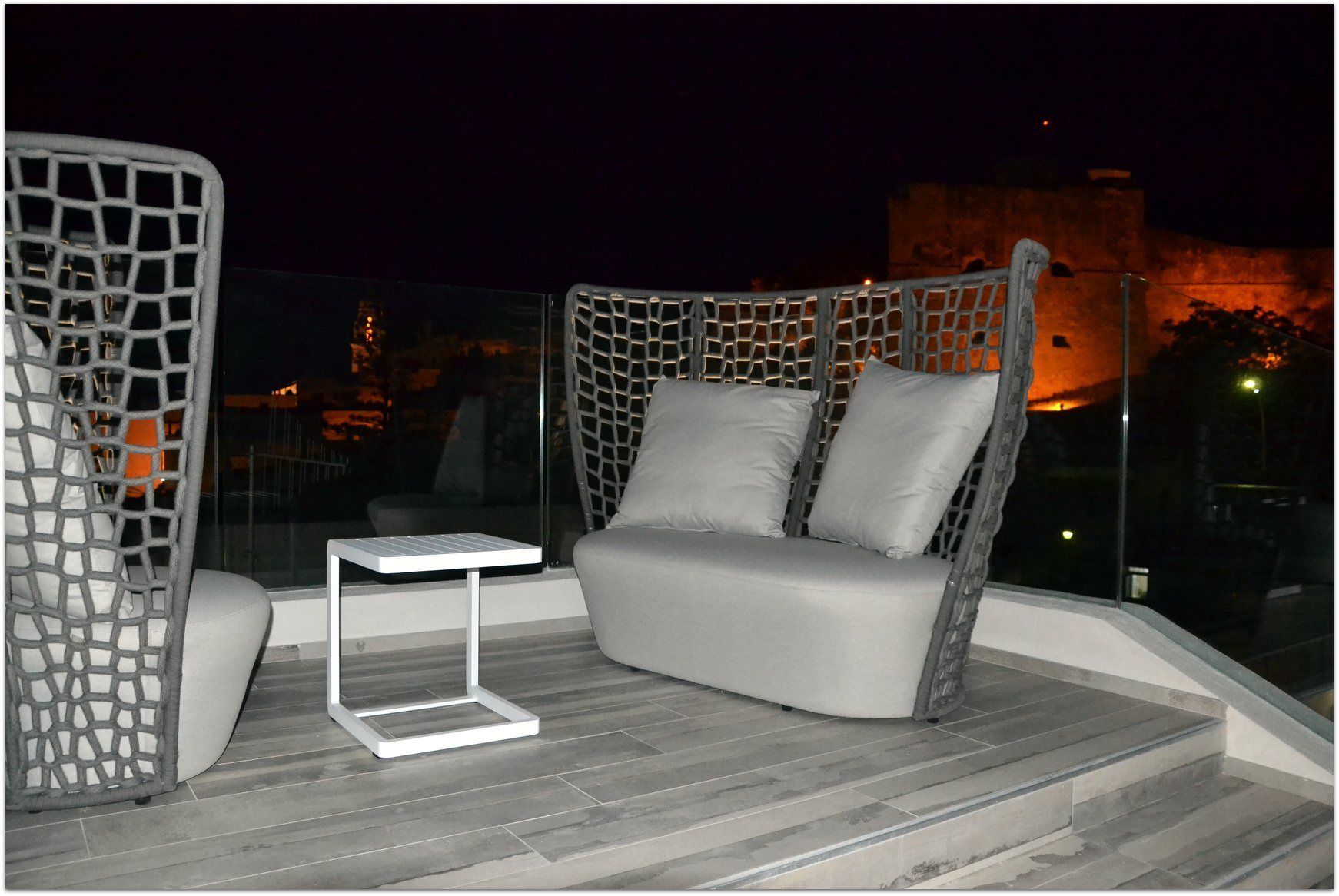 Un balcón con zona de estar acolchada, una mesita y vistas a un paisaje urbano nocturno.