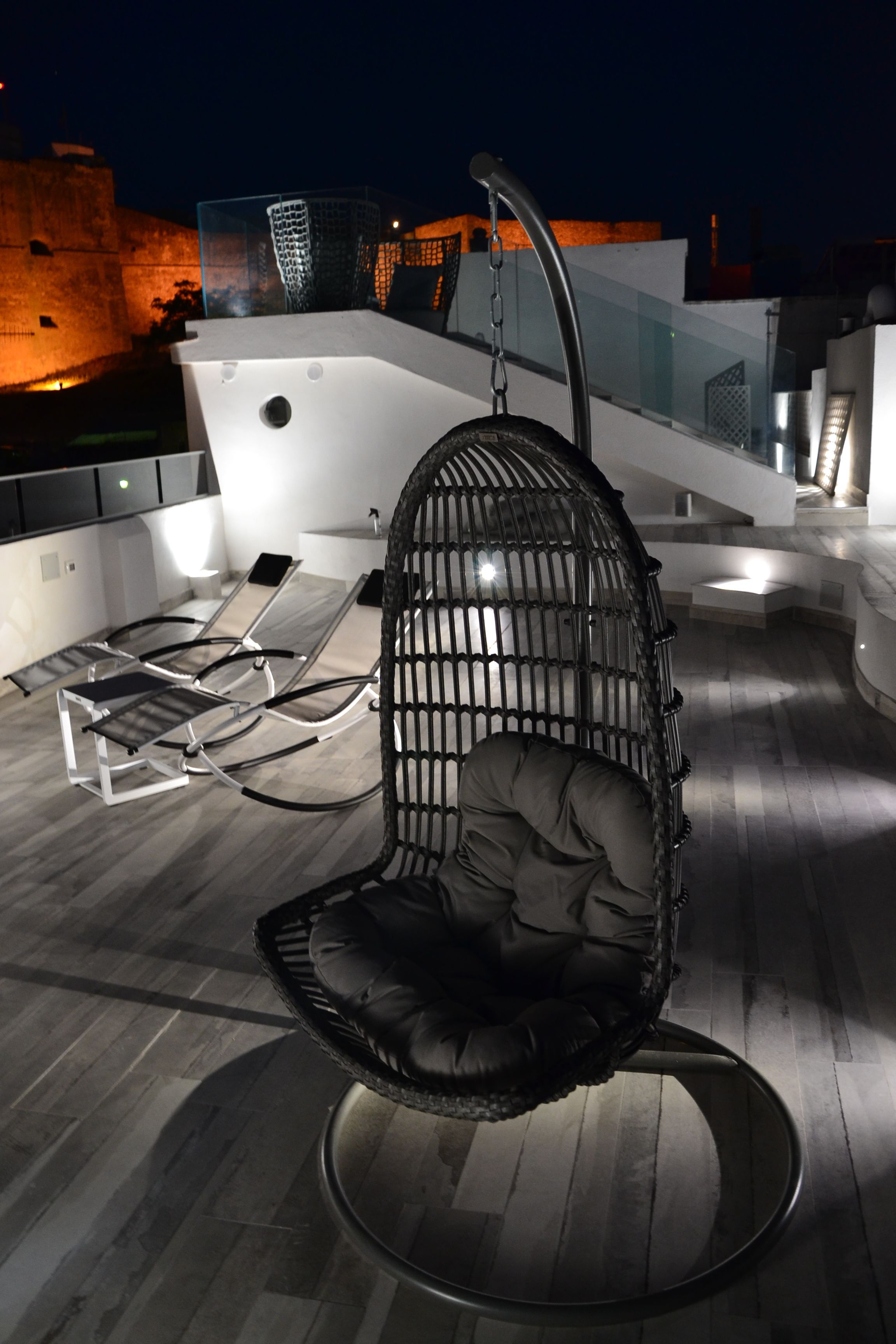 A modern outdoor seating area with a hanging chair and lounge chairs, a staircase, and a view of a building in the background.