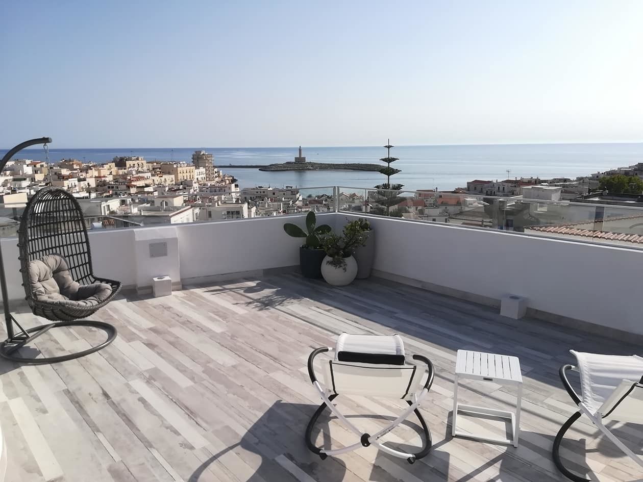 A rooftop terrace with a view of the sea, a hanging chair, two lounge chairs, and potted plants.