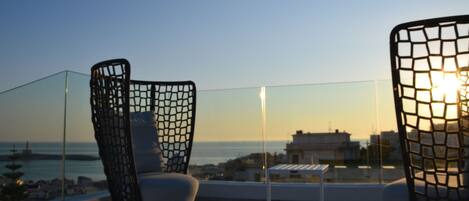 Uma varanda com duas cadeiras, uma mesa pequena e vista para o mar e edifícios ao pôr do sol.