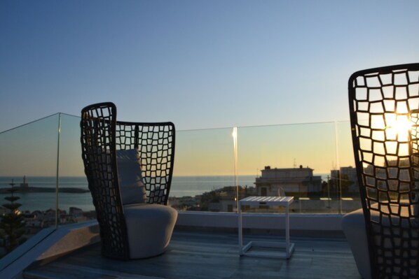 A balcony with two chairs, a small table, and a view of the sea and buildings during sunset. - Quintessenza - Charme Rooms (Vieste)