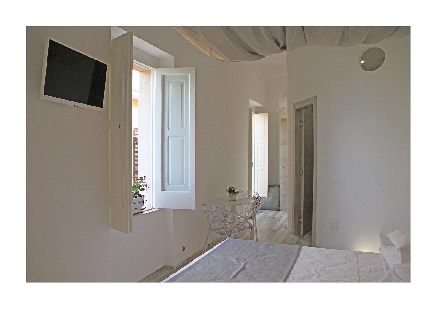 Un dormitorio con cama, televisor montado en la pared, ventana y puerta que da a otra habitación.