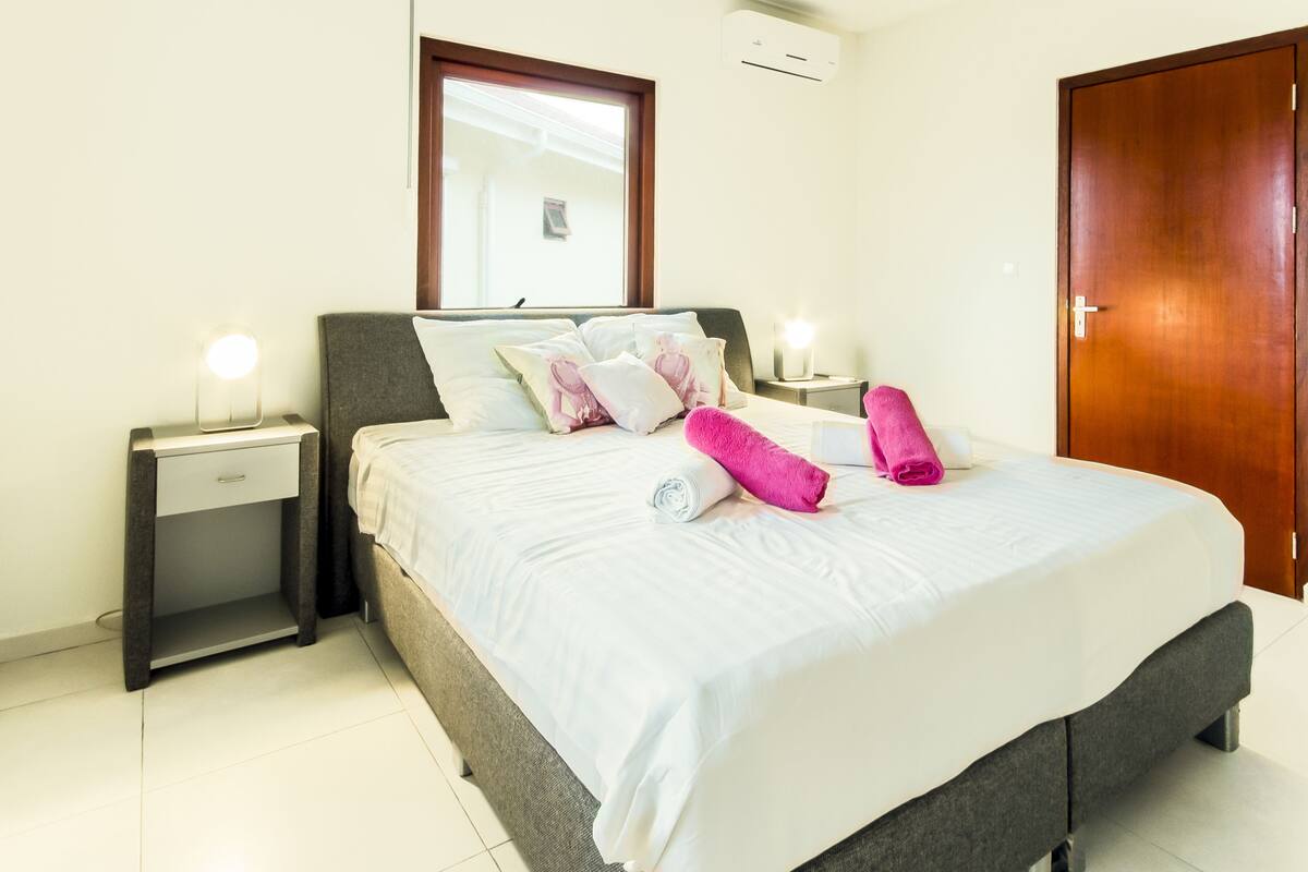 A neatly made bed with white linens and pink towels, a bedside table with a lamp, a nightstand, a wooden door, and a window with blinds.