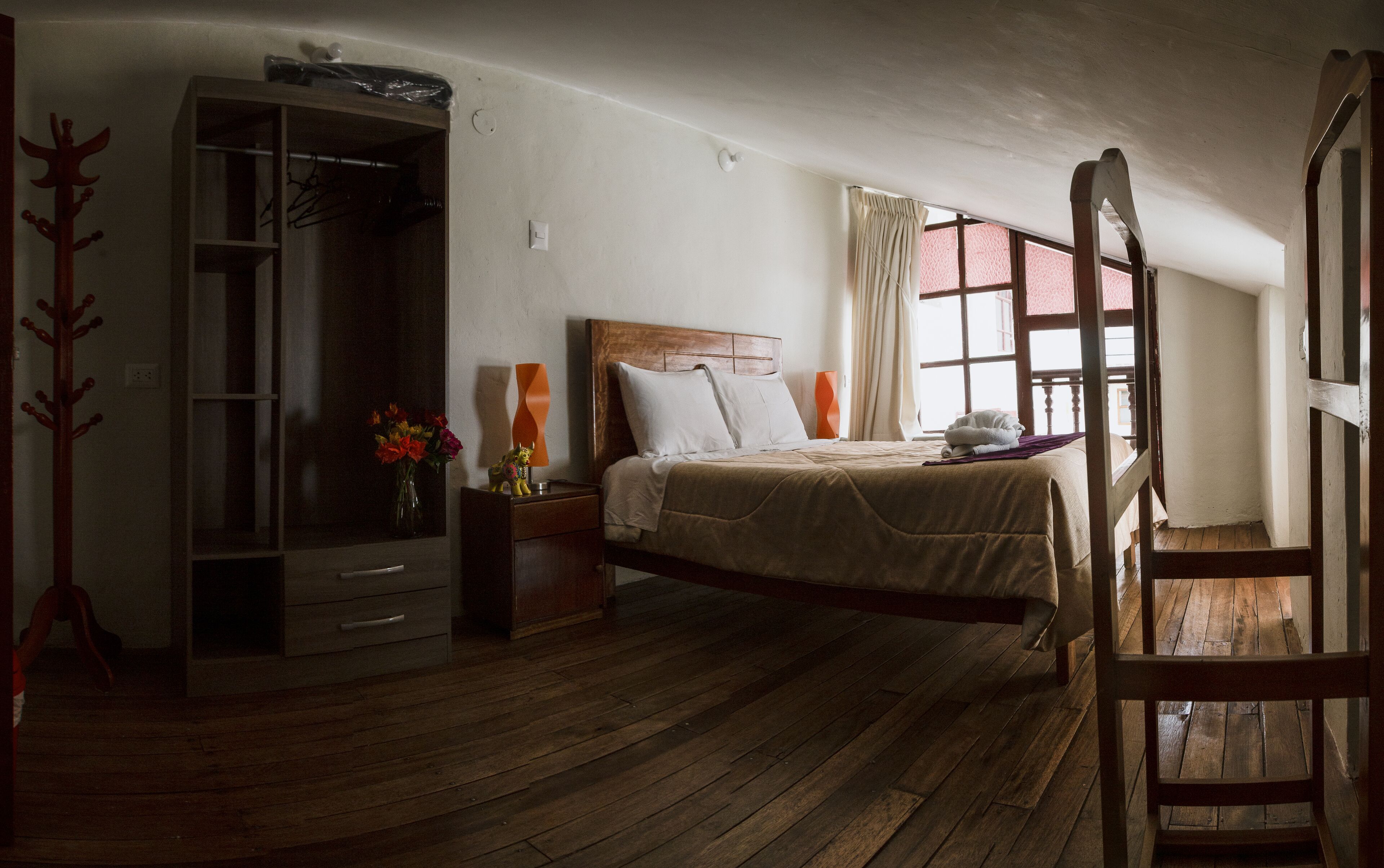 Un dormitorio con piso de madera, una cama con cabecera de madera, una mesita de noche, un aparador, un espejo y una ventana con cortinas.