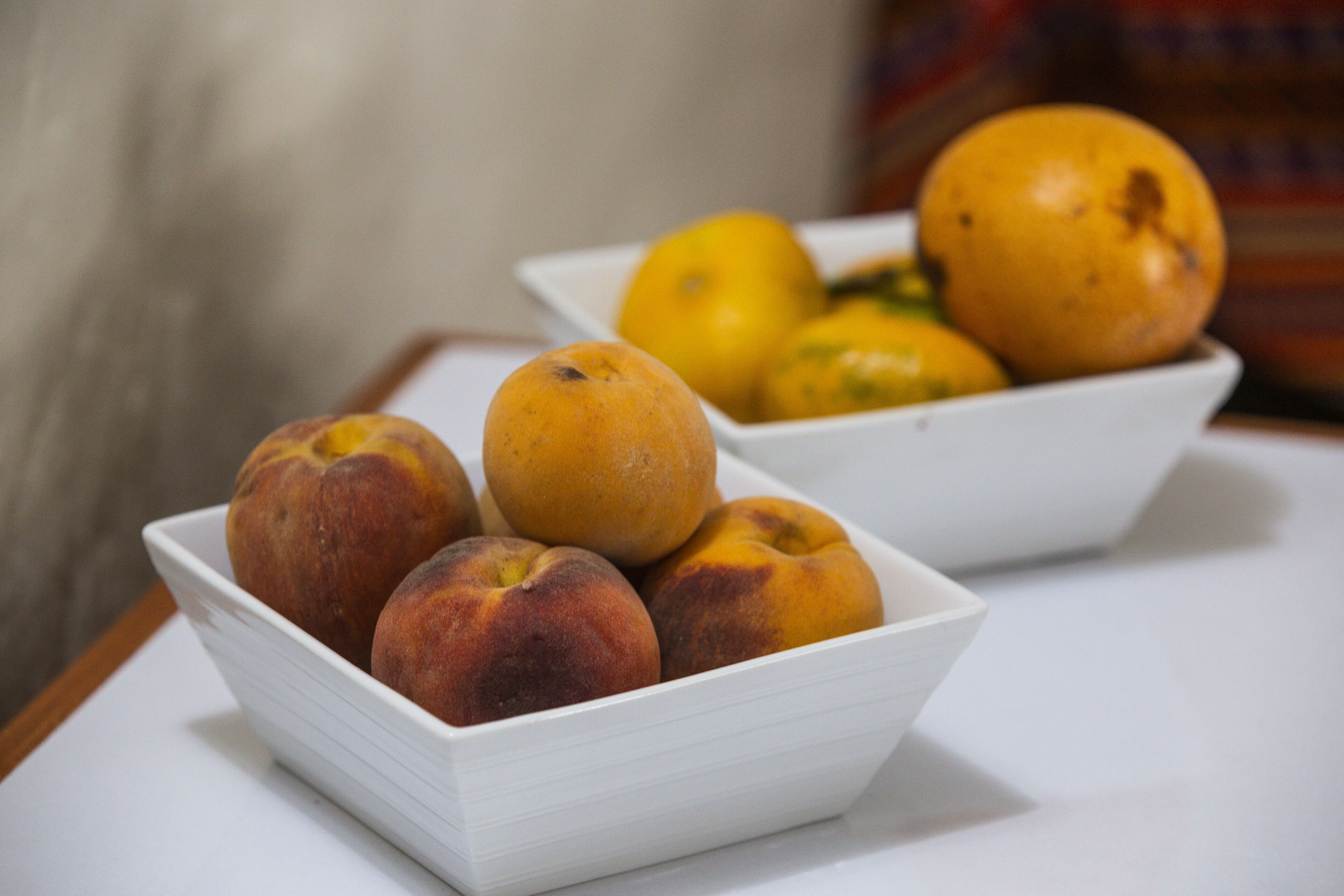 Two white bowls containing peaches and oranges on a table.
