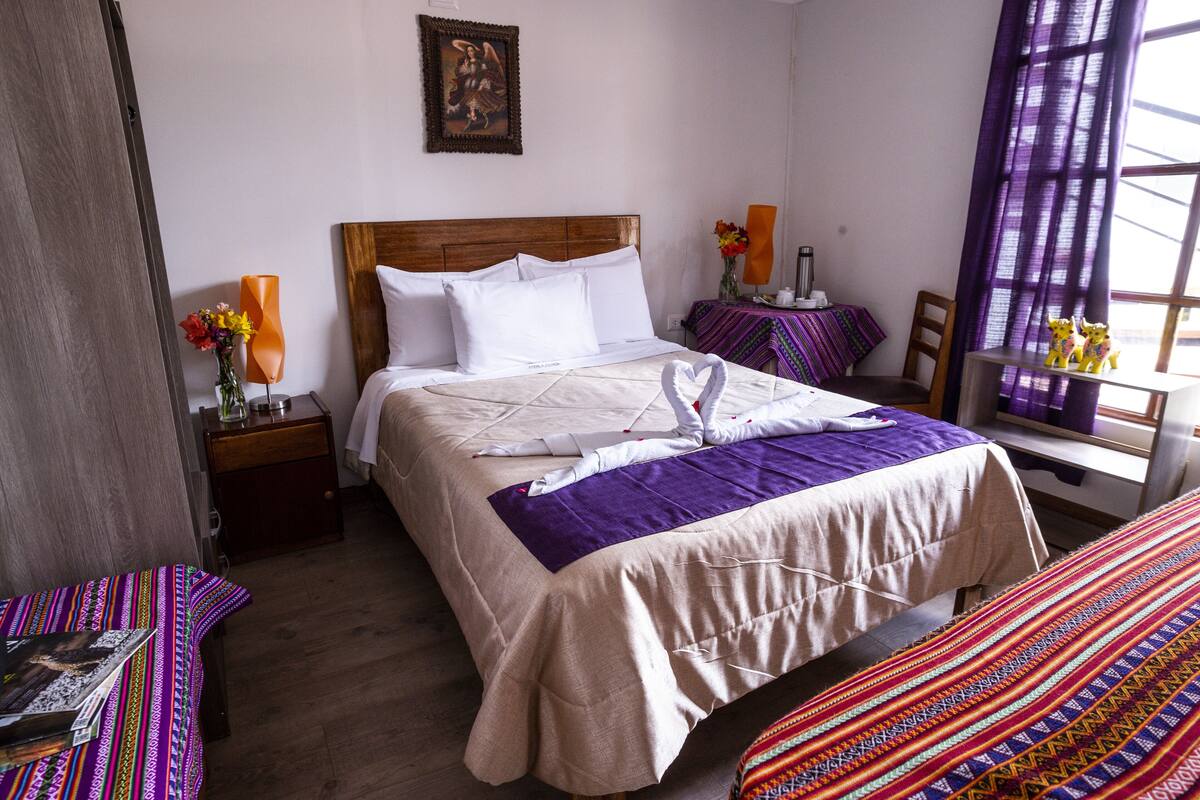 Ein Hotelzimmer mit einem großen Bett, einem Holz-Kopfstück, einem Nachttisch mit Lampe, einem kleinen Tisch mit Blumenvase und einem Fenster mit violetten Vorhängen.