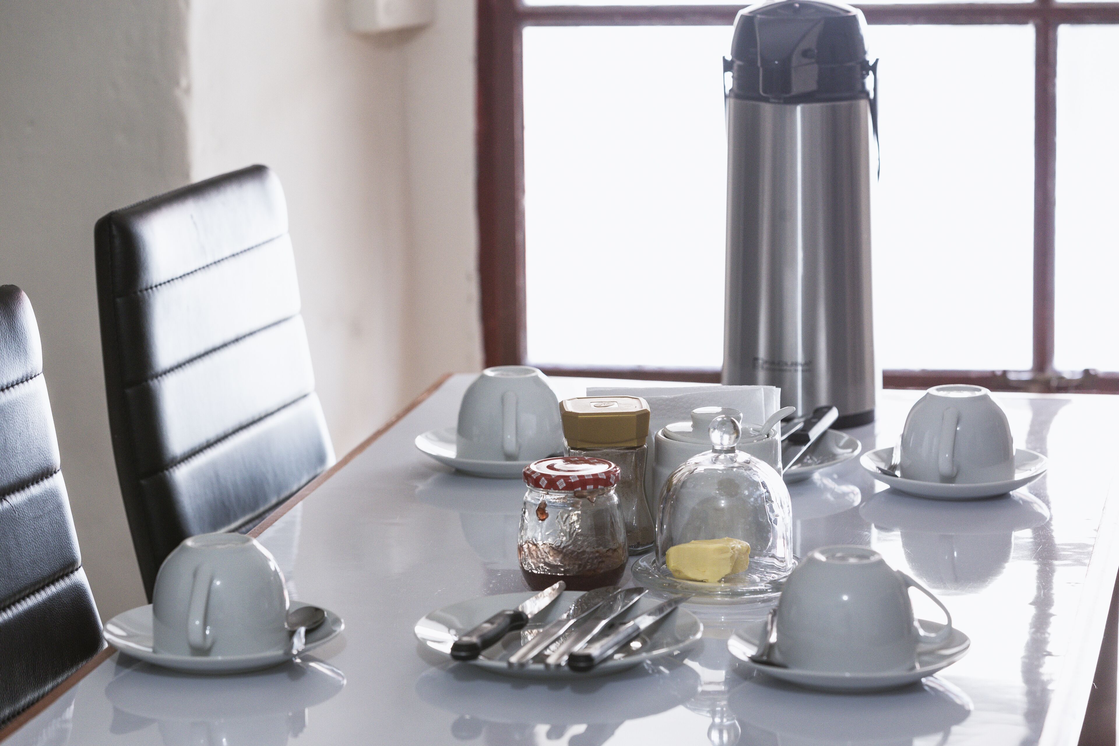 A dining table with white cups, saucers, a thermos, a jar of jam, a butter dish, and cutlery.