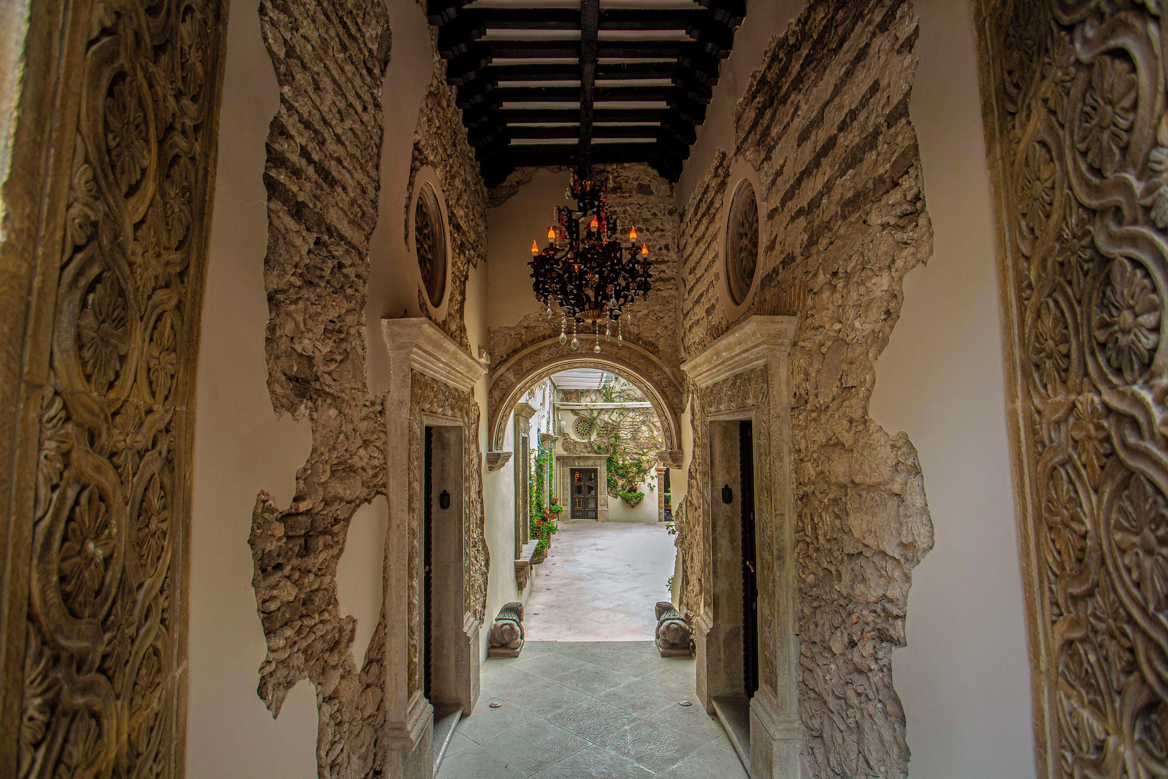 An ornate hallway with a chandelier, stone walls, and an archway leading to a courtyard.