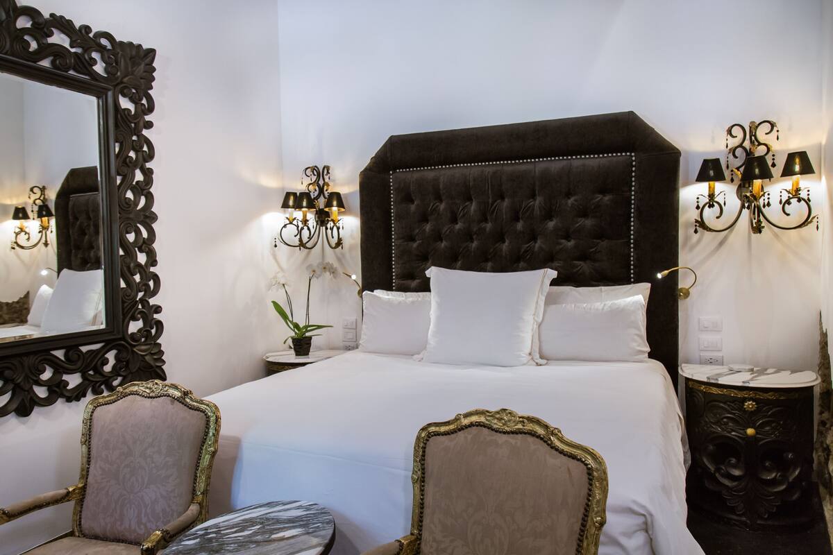Un grand lit doté d’une tête de lit capitonnée, deux tables de chevet avec des lampes, un miroir avec un cadre orné et deux fauteuils rembourrés.