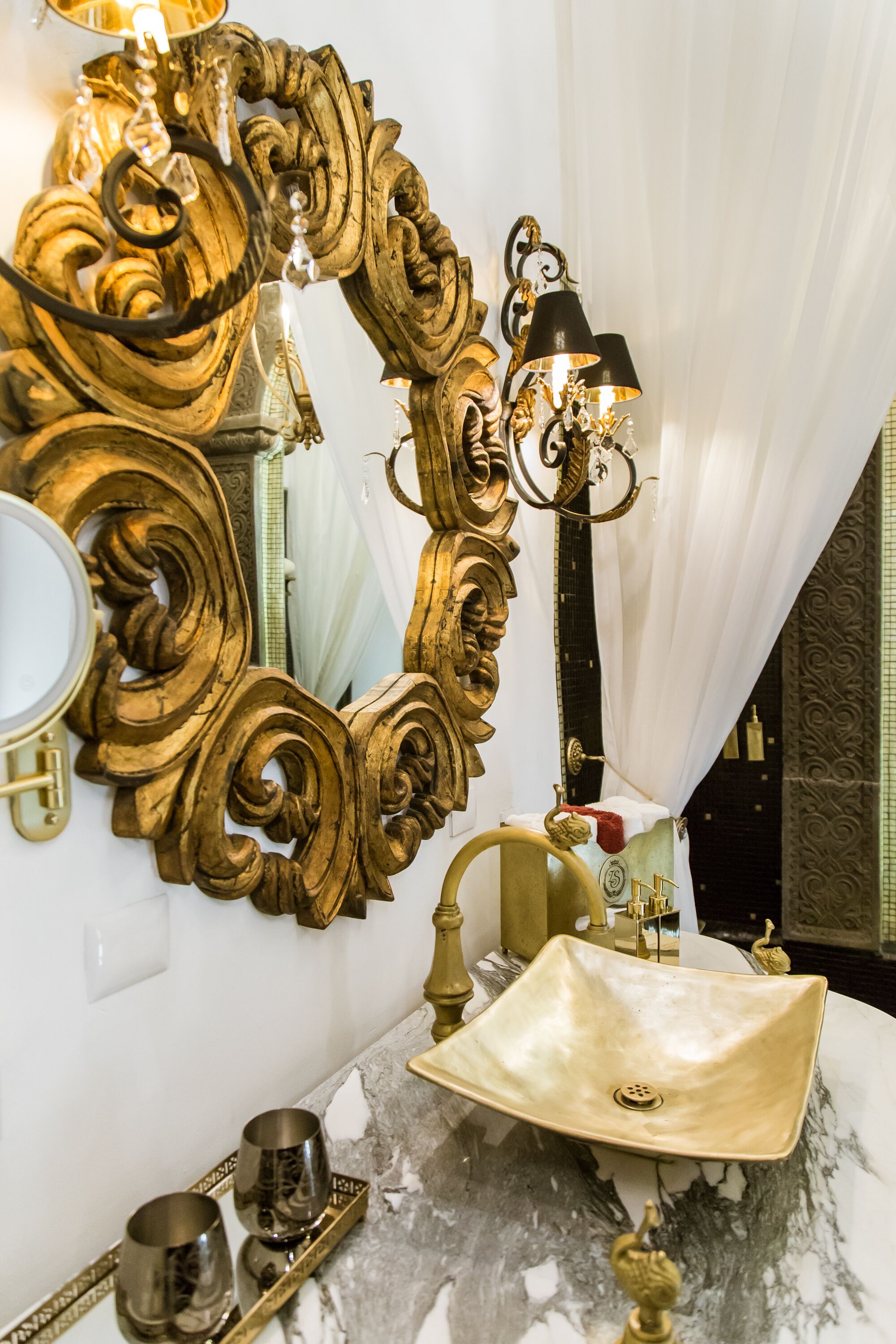 Un baño con un espejo grande y ornamentado, un lavamanos dorado y una lámpara de pared.