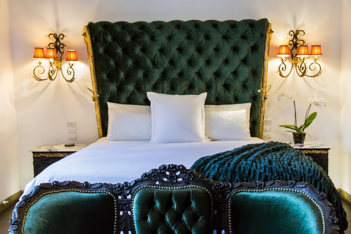 Un grand lit doté d’une tête de lit capitonnée, de draps blancs et d’un plaid vert foncé. Deux tables de chevet, sur lesquelles sont posées des lampes et une plante en pot sur l’une d’elles, se trouvent de chaque côté du lit.