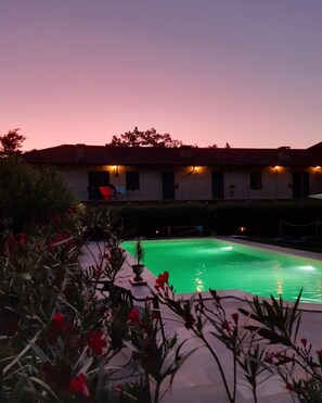 A swimming pool with a tiled deck, surrounded by greenery and a building with multiple windows and a red roof. - La Cascina del Castello (Asti)
