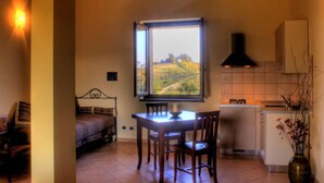A room with a bed, a dining table with chairs, a window with a view of a landscape, a kitchen area with a stove and cabinets, and a vase with flowers. - La Cascina del Castello (Asti)